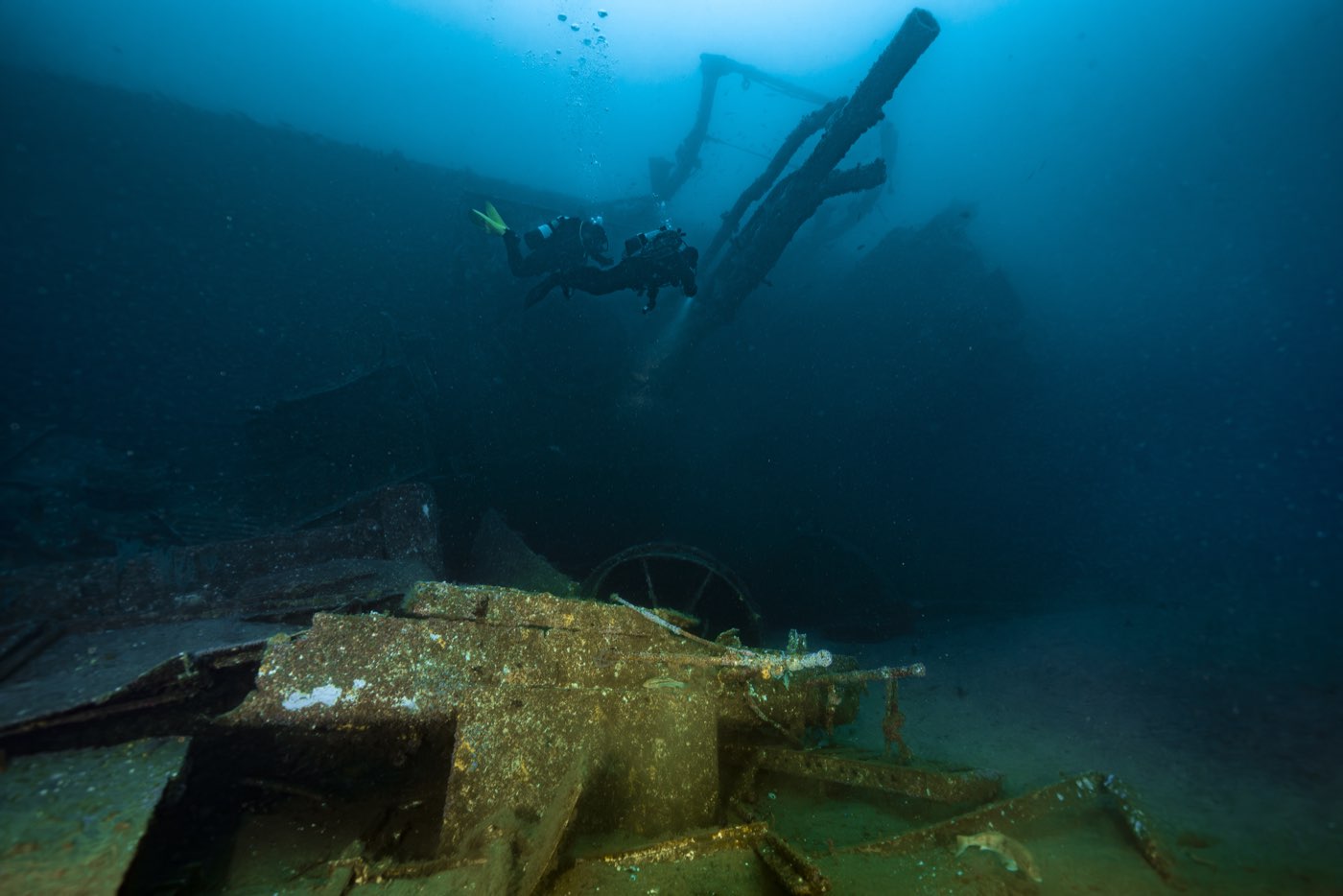 The smokestack debris field on the sand beside the hull, two divers passing over it